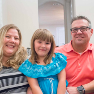 Family portrait featuring a smiling woman in a striped shirt, a young girl in a blue dress, and a man in a pink shirt, all sitting together in a cozy indoor setting, reflecting the friendly and supportive atmosphere of Zoom Casa's services.
