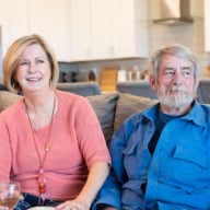 Smiling couple sitting together on a couch in a modern living room, representing satisfied Zoom Casa clients sharing a positive home-selling experience.