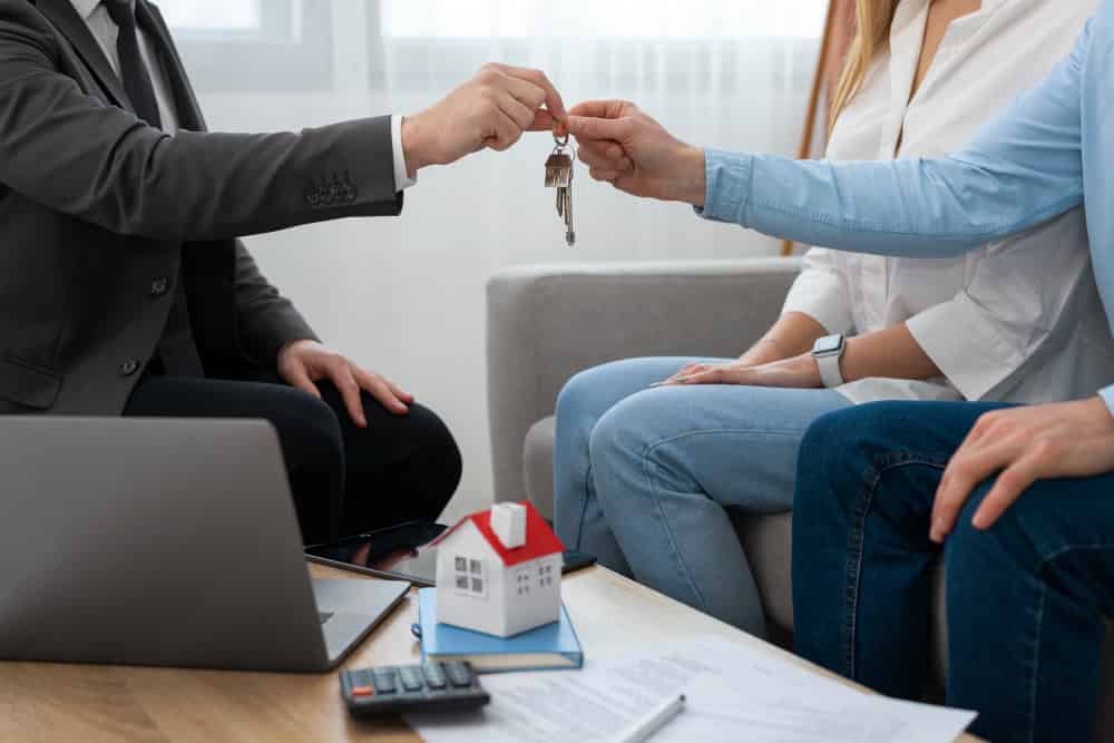 Real estate agent handing house keys to buyers, with a miniature house model and paperwork on the table, representing home selling process and client engagement.