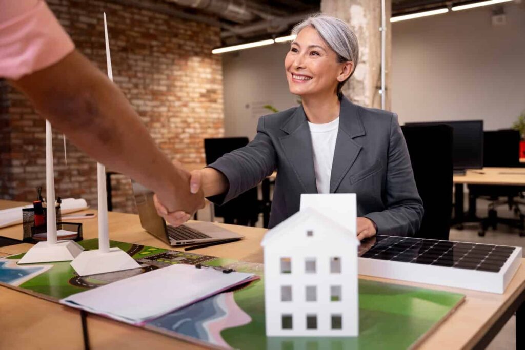 Business professional shaking hands over a table with a small model house, solar panel, and architectural plans, symbolizing collaboration in real estate and alternative energy solutions.
