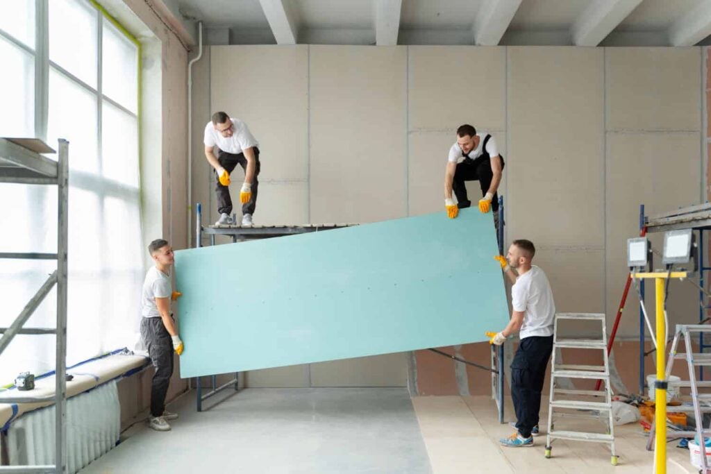 Four workers collaborating to lift and install a large sheet of drywall in a partially renovated room, emphasizing teamwork in home renovation for enhanced property value.