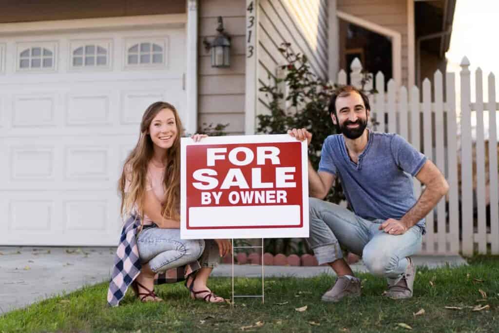 Couple smiling and posing with a "For Sale By Owner" sign in front of their home, showcasing the selling process and enhancing curb appeal.