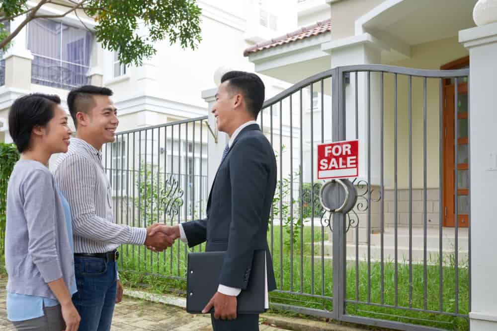Couple shaking hands with real estate agent in front of a house for sale, highlighting home selling process and buyer engagement with Zoom Casa services.