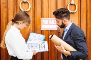 Man and woman discussing home selling strategies in front of a wooden door with a "For Sale" sign, holding property plans and documents.