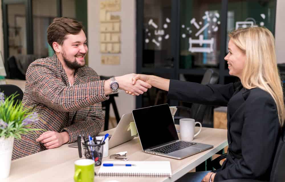 Man and woman shaking hands at a desk with laptops and coffee cups, representing successful real estate negotiations and client relationships.