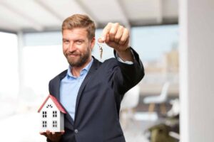 Man in a suit holding a small house model and keys, symbolizing home sales and real estate success.